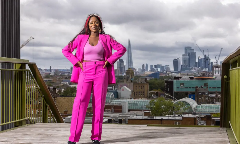 A woman in a bright pink suit, smiling and standing in a confident pose on a rooftop with a dramatic, cloudy London skyline in the background
