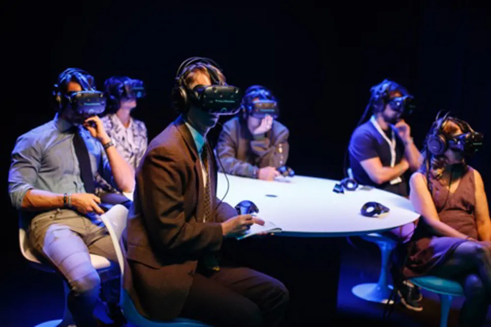 A group of 6 people sitting at a table wearing virtual reality headsets.