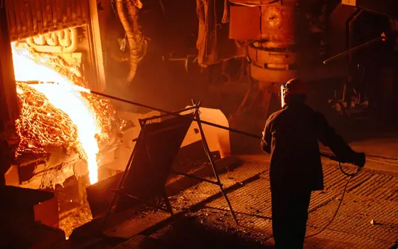 A worker wearing protective gear is standing several metres away from the outlet of a blast furnace, pushing a temperature probe into the white-hot molten metal pouring from it.