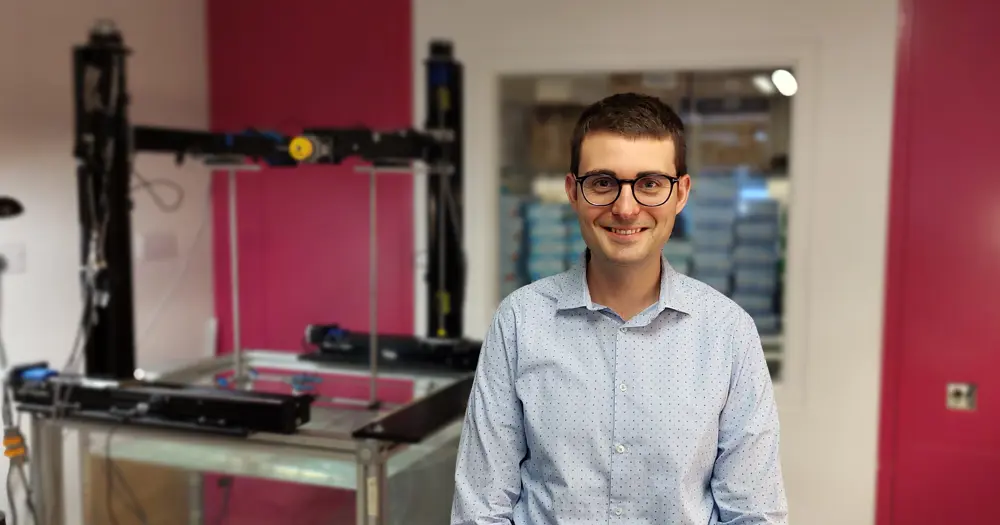 Oscar Calderon Agudo in a lab‑style workspace with a large piece of industrial equipment behind him, including metal framing and mechanical components.