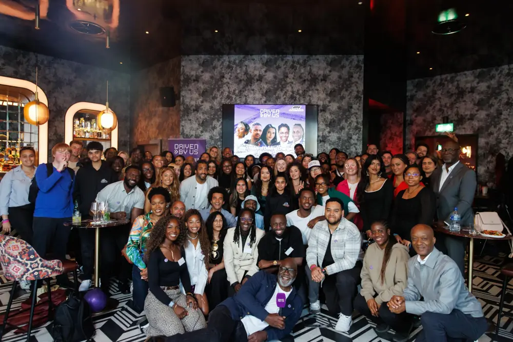 A large, diverse group of people pose together at a social event in a bar, with a screen behind them reading ‘Driven by us’.