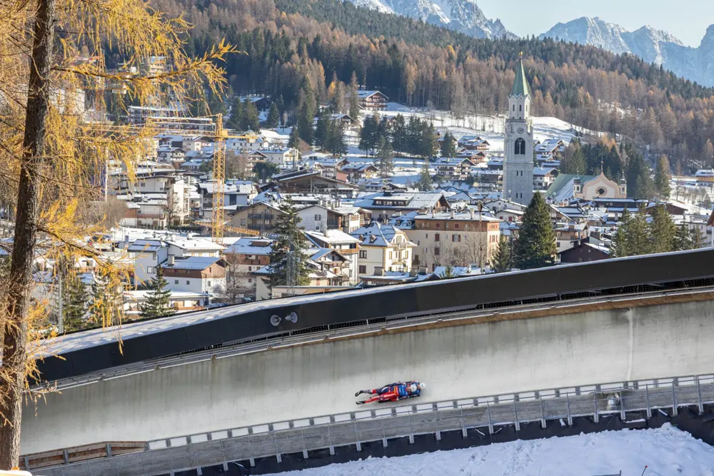 Two bobsledders speed around a curved ice track above snowy Cortina D'Ampezzo, with mountains rising behind the buildings.
