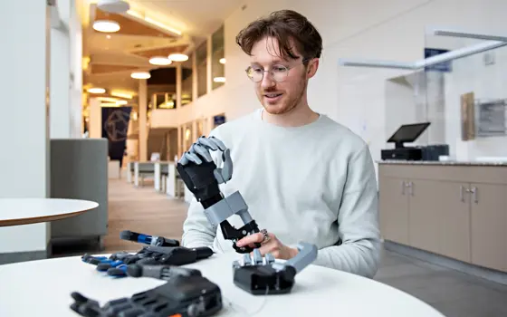 A man sitting at a table, holding a prototype of a prosthetic hand based on the classic 'split hook' style