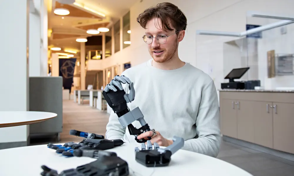 A man sitting at a table, holding a prototype of a prosthetic hand based on the classic 'split hook' style