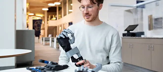 A man sitting at a table, holding a prototype of a prosthetic hand based on the classic 'split hook' style