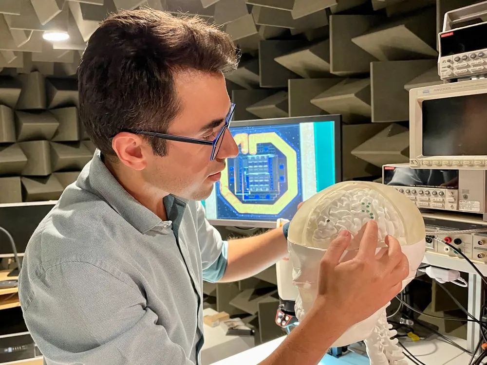 A man with brown hair and glasses holds a model of a human brain, with six small green circles positioned on the surface, perhaps indicating where MintNeuro's implants would be placed.