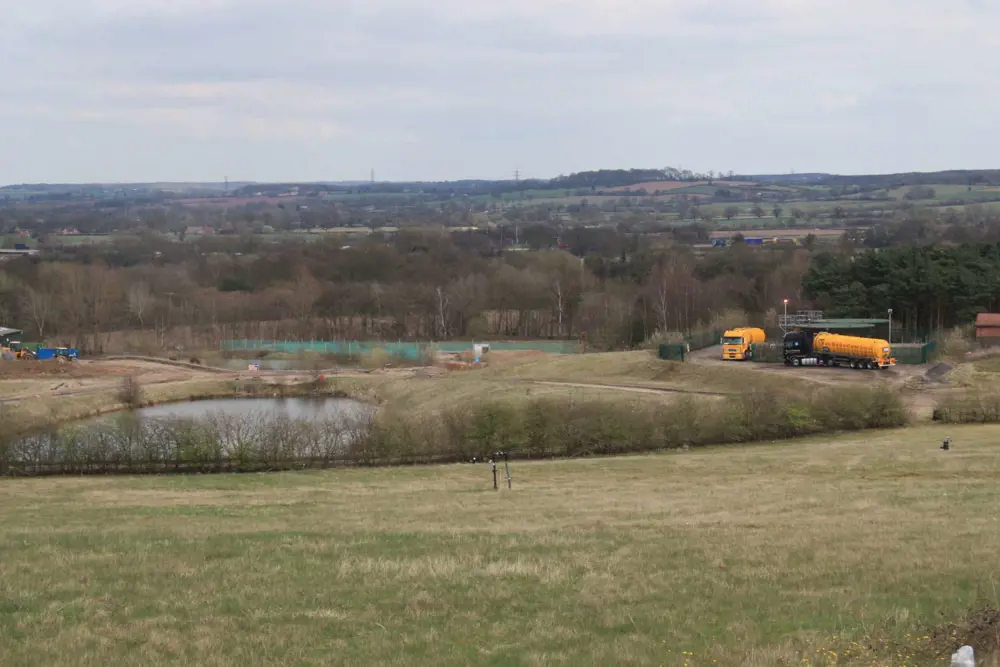 A field, which sits on top of an old landfill site, with a lake, trees and two large lorries in the background.