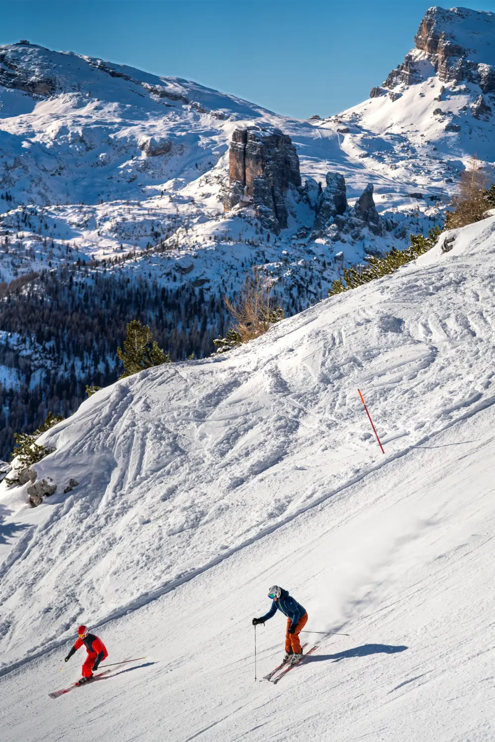 Two skiers descend a sunny mountain slope with rugged, snow‑covered peaks in the background.