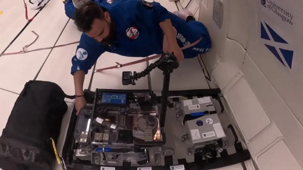 A man on a hyperbolic flight wearing a blue boiler suit, floating off the ground as he adjusts his 3D printer.