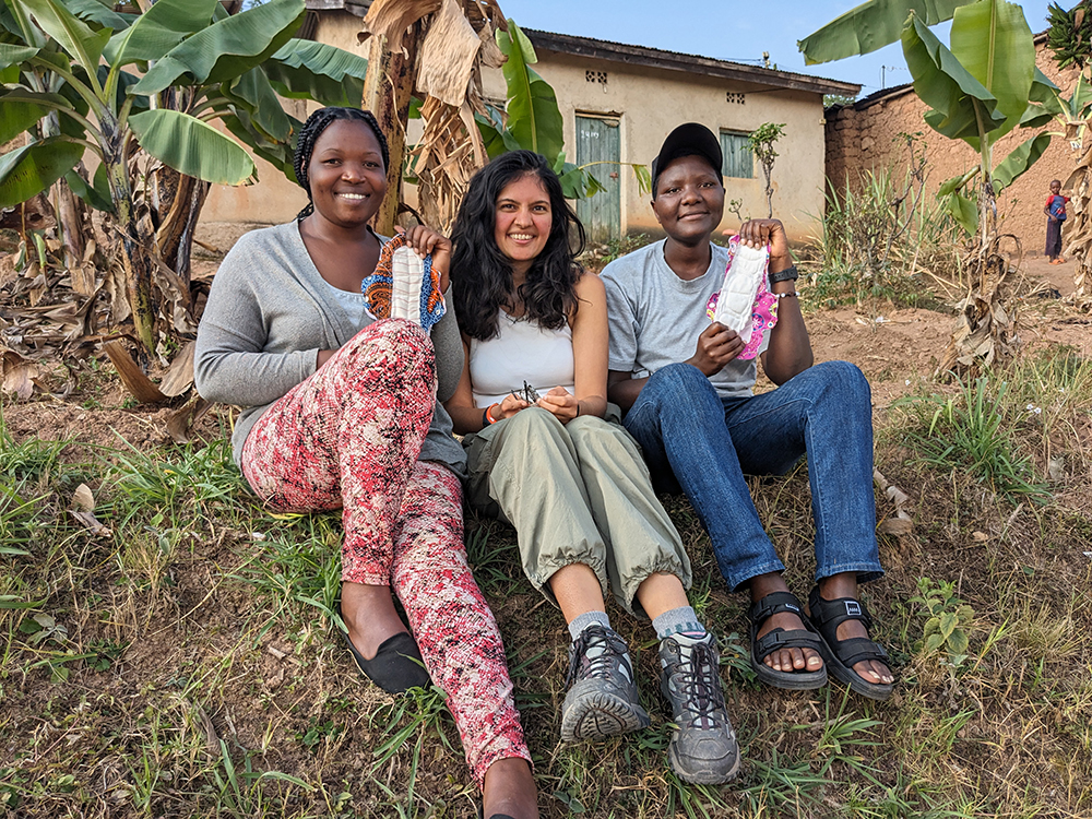 Three people sit on grass in a rural setting, holding reusable fabric pads, with banana plants and small houses in the background.