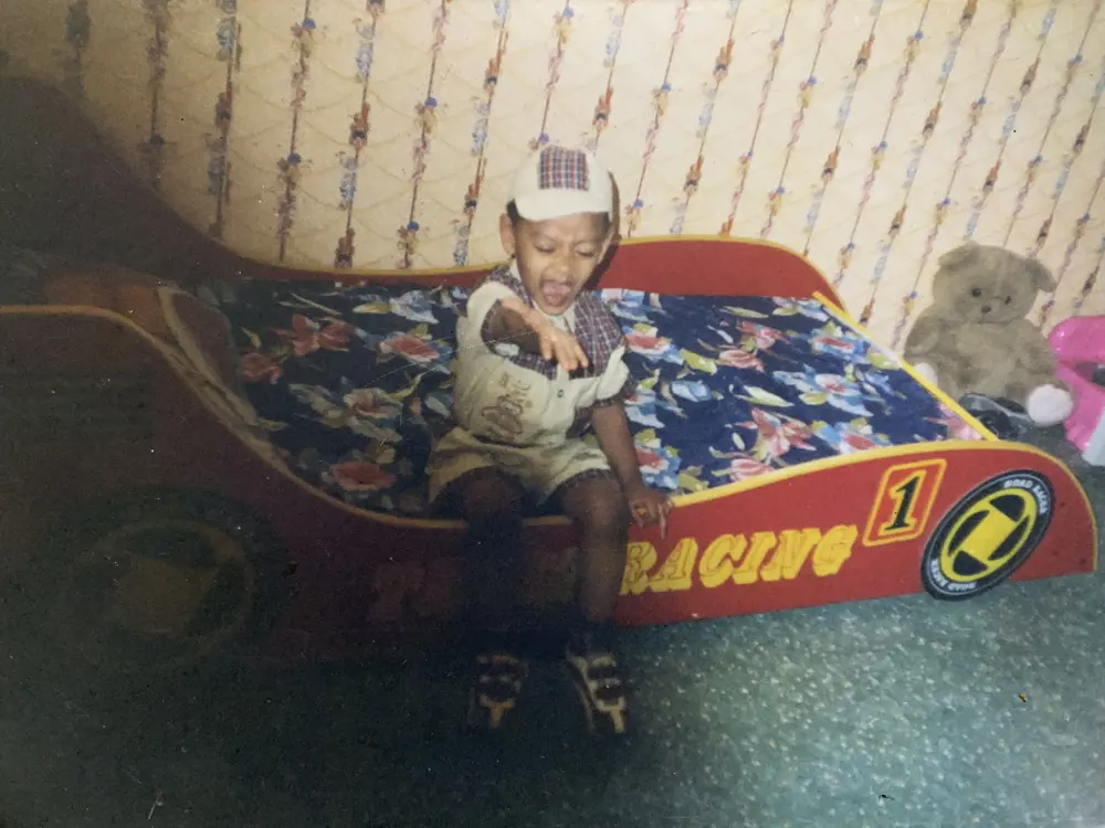 A young boy wearing a cap, sitting on a bed shaped like a racecar and gesturing at the camera