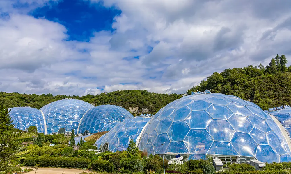 Large transparent geodesic domes of the Eden Project sit among green hills under a bright, partly cloudy sky.