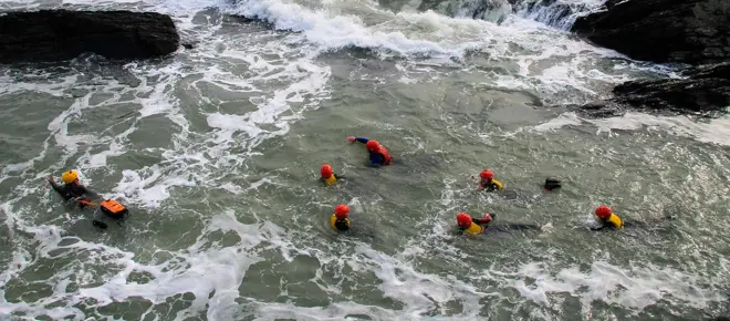 A group of seven "coasteerers" swimming around a rocky section of a coastline, all wearing red helmets and yellow lifejackets