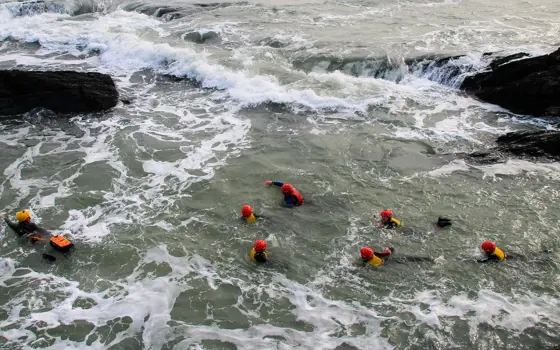 A group of seven "coasteerers" swimming around a rocky section of a coastline, all wearing red helmets and yellow lifejackets
