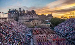 A wide view of the Edinburgh Castle Esplanade at sunset, with full grandstands on both sides and massed performers in military uniforms assembled in formation on the parade ground during the Royal Edinburgh Military Tattoo.