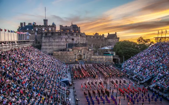 A wide view of the Edinburgh Castle Esplanade at sunset, with full grandstands on both sides and massed performers in military uniforms assembled in formation on the parade ground during the Royal Edinburgh Military Tattoo.