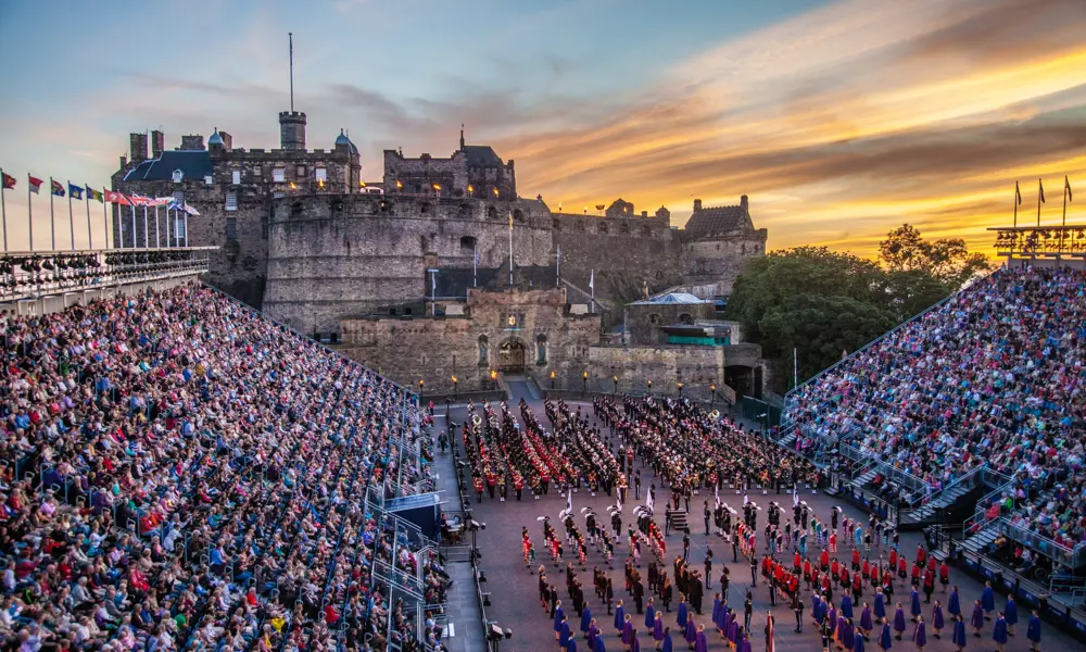 A wide view of the Edinburgh Castle Esplanade at sunset, with full grandstands on both sides and massed performers in military uniforms assembled in formation on the parade ground during the Royal Edinburgh Military Tattoo.