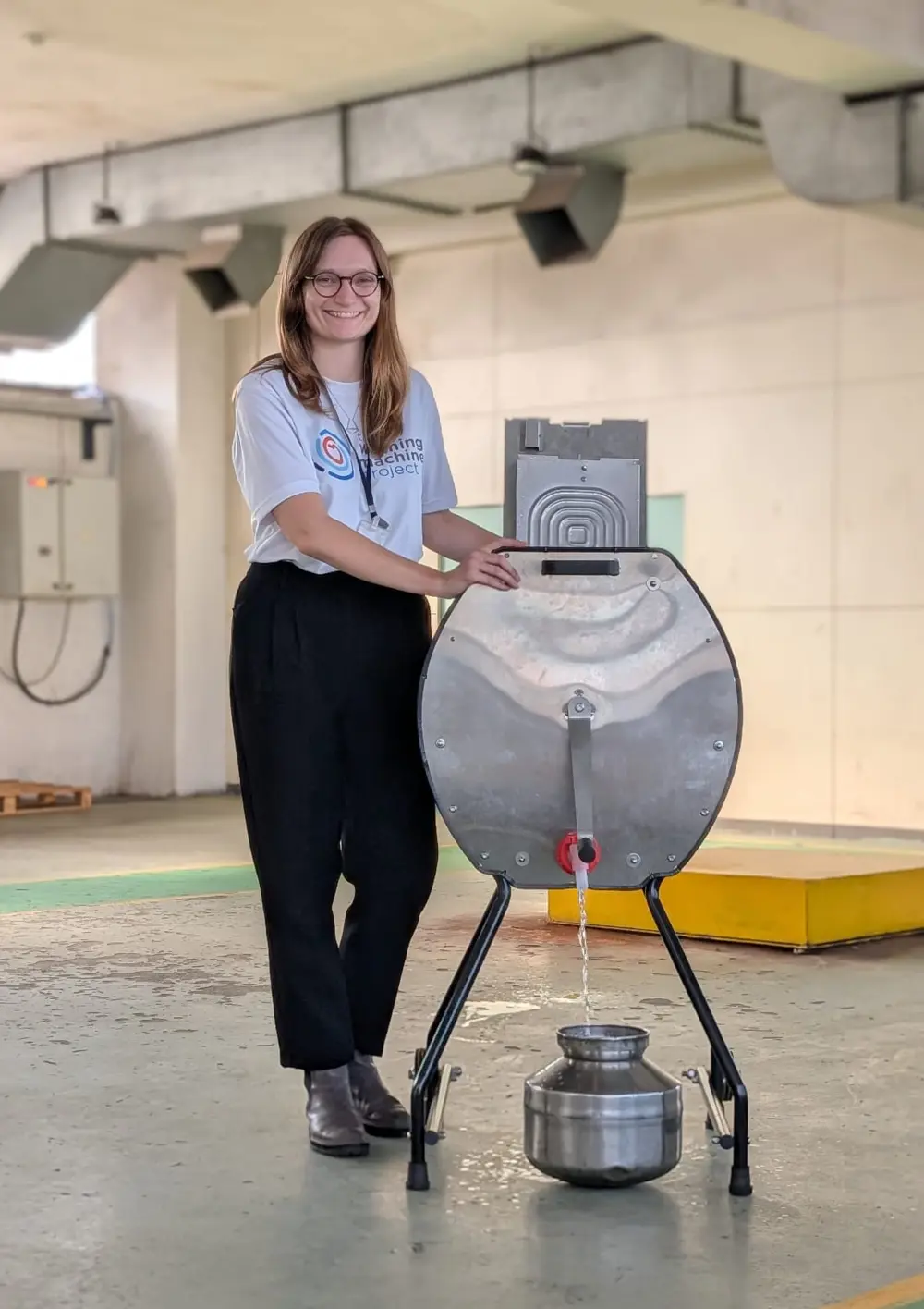 Person standing beside a metal device on a stand, with liquid flowing into a container in an indoor workshop setting. 