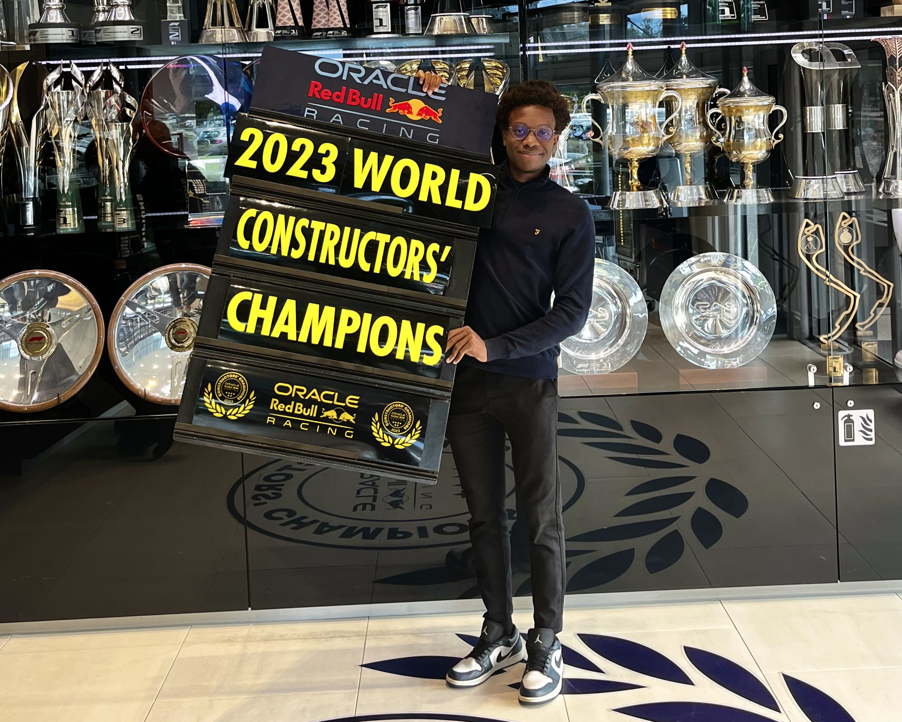 A man smiles for a photo, standing in front of a trophy cabinet and holding a sign that reads "2023 World Constructors' Championship", which shows the Red Bull Racing and Oracle logos.