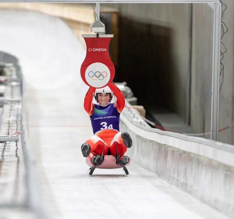 A luge athlete races down the Eugenio Monti ice track at Cortina D'Ampezzo during testing sessions for the 2026 Winter Olympics, passing under a timing banner displaying the Olympic rings.