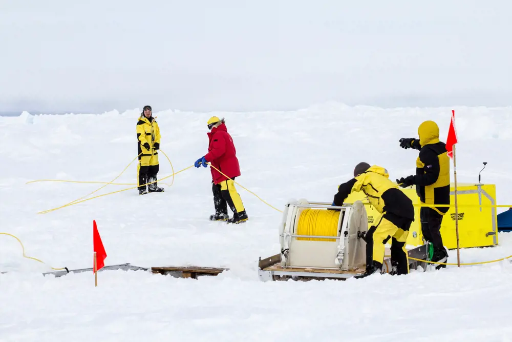 People wearing bright yellow, red and black snowsuits uncoiling a yellow cable on a snowy landscape.