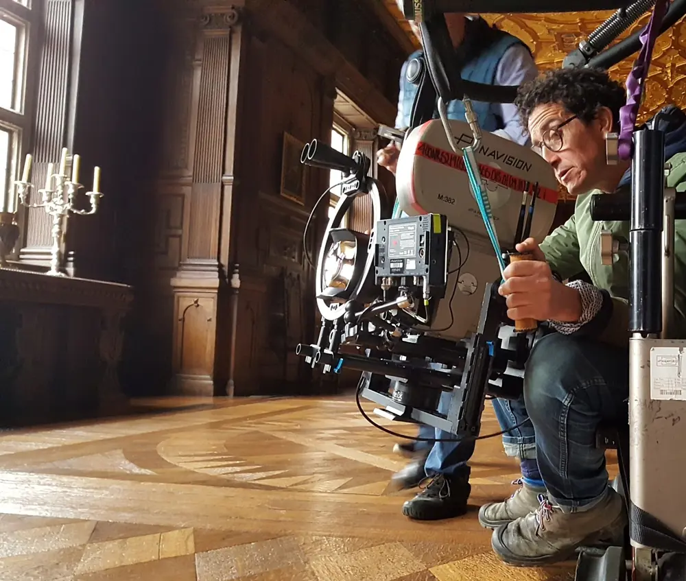 A professional film camera mounted on a stabilising rig being adjusted by a crew member in an elaborate interior with carved wooden walls and decorative flooring.