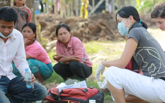 Dr Anh Tran crouching down with a mask and gloves on, with chemical bottles in front of her on the ground, being watched by local people in Cambodia.