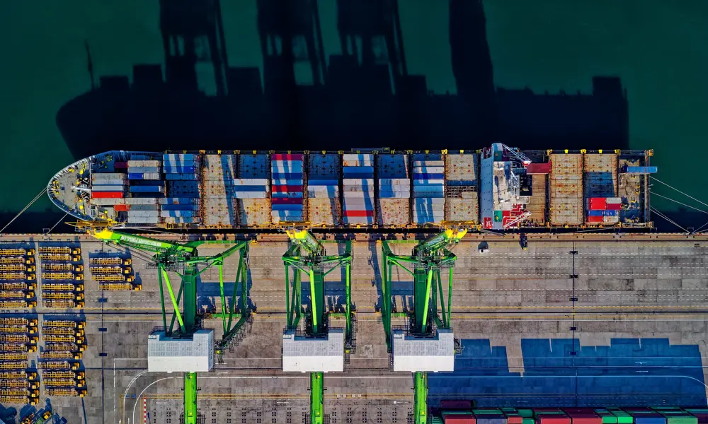 A container ship viewed from above, with green cranes on the dock.