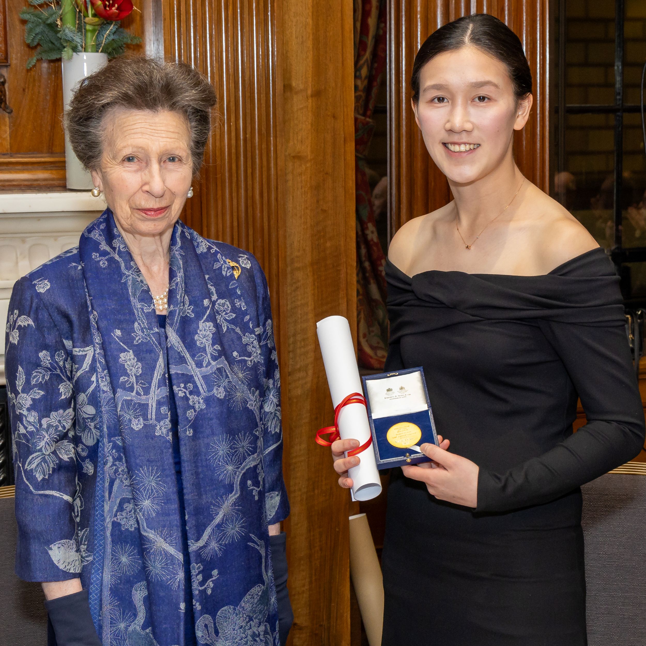 Princess Anne and Rachel Chiu are in evening wear, standing indoors in a wood-panelled room; Rachel holds a certificate scroll and an open presentation case with a medal, indicating a formal award ceremony.