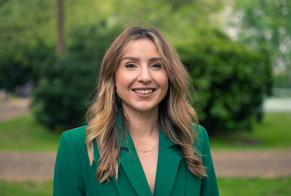 Portrait of a person in a green blazer standing outdoors, with trees and a park pathway in the background.
