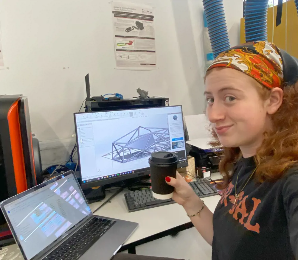 A girl with red hair wearing a bandana, holding a takeaway coffee and sitting at a desk in front of a laptop and desktop computer with an engineering design on the screen.