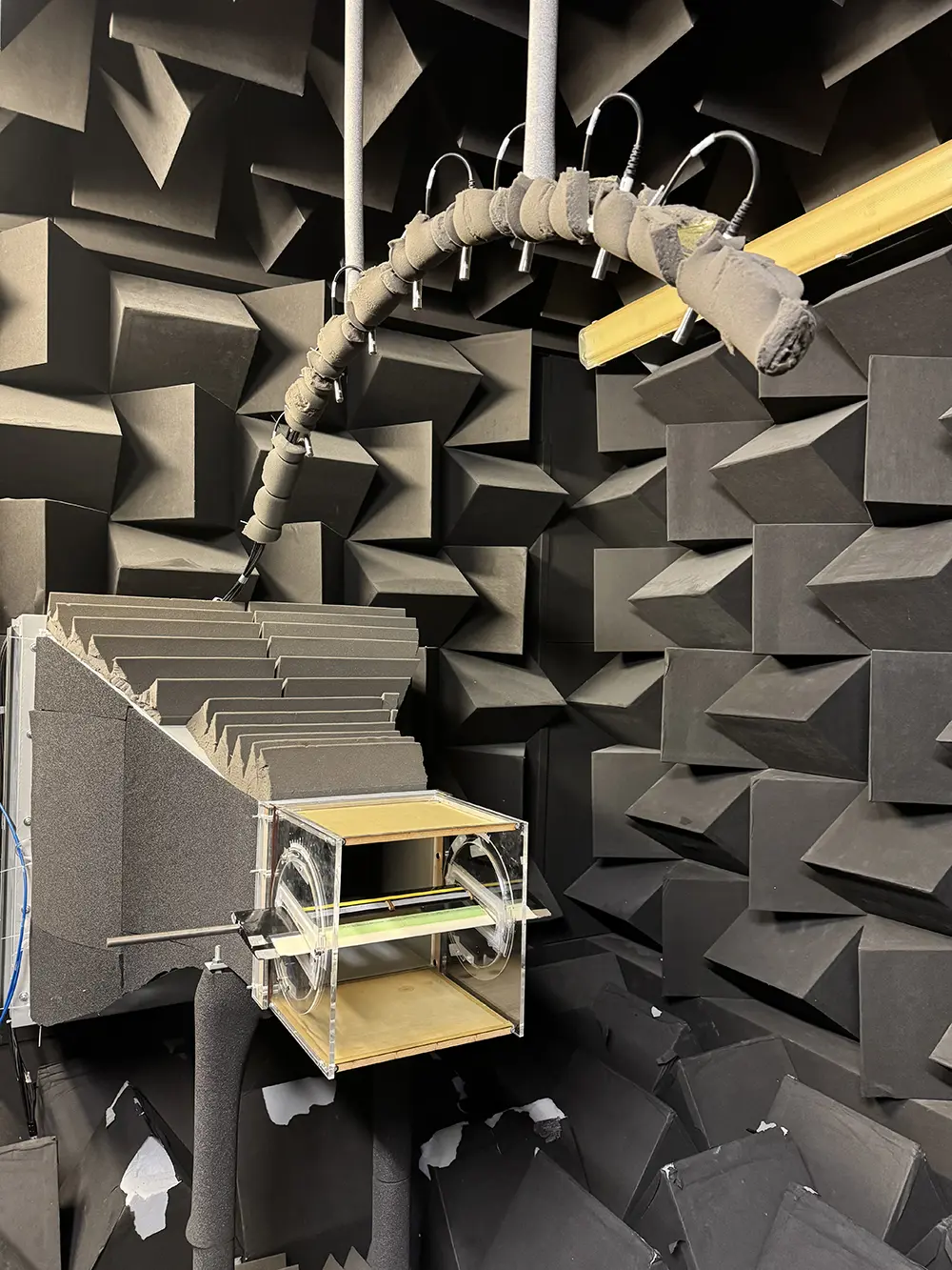 A small-scale model of an aeroplane wing is held in a box with two wooden sides and two clear sides that makes up a wind tunnel. It is in a grey soundproof room with a microphone arched over the top. 