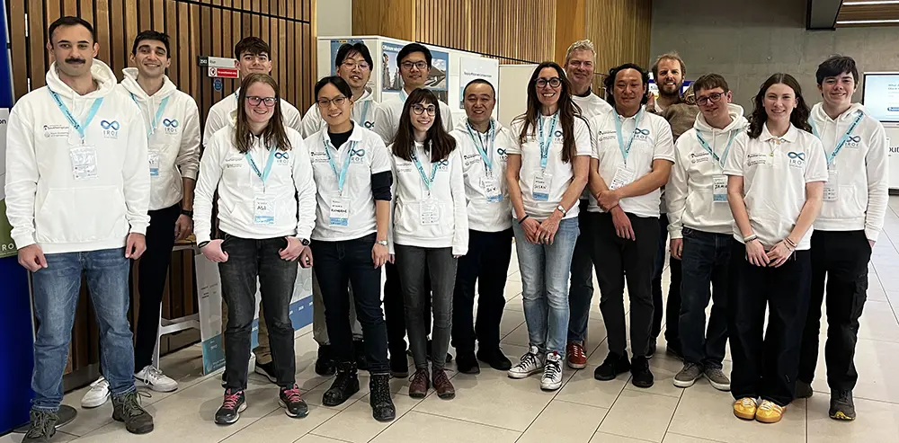 A group of people all wearing white t-shirts and lanyards stand together in a conference hall, smiling at the camera