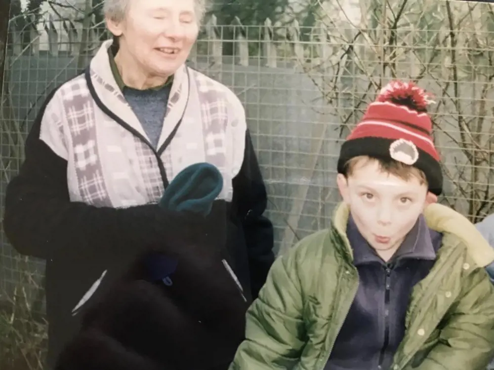 A young boy wearing a coat and a bobble hat, pulling a comedic face and standing with his grandmother, who is laughing