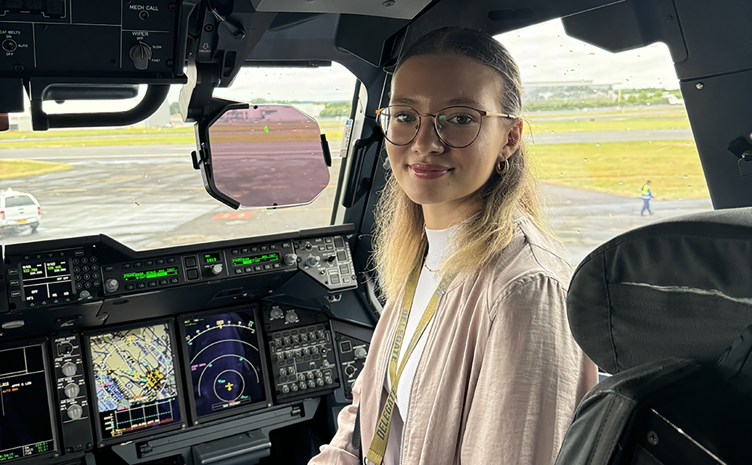 A blond woman wearing glasses sits inside a cockpit, looking at the camera
