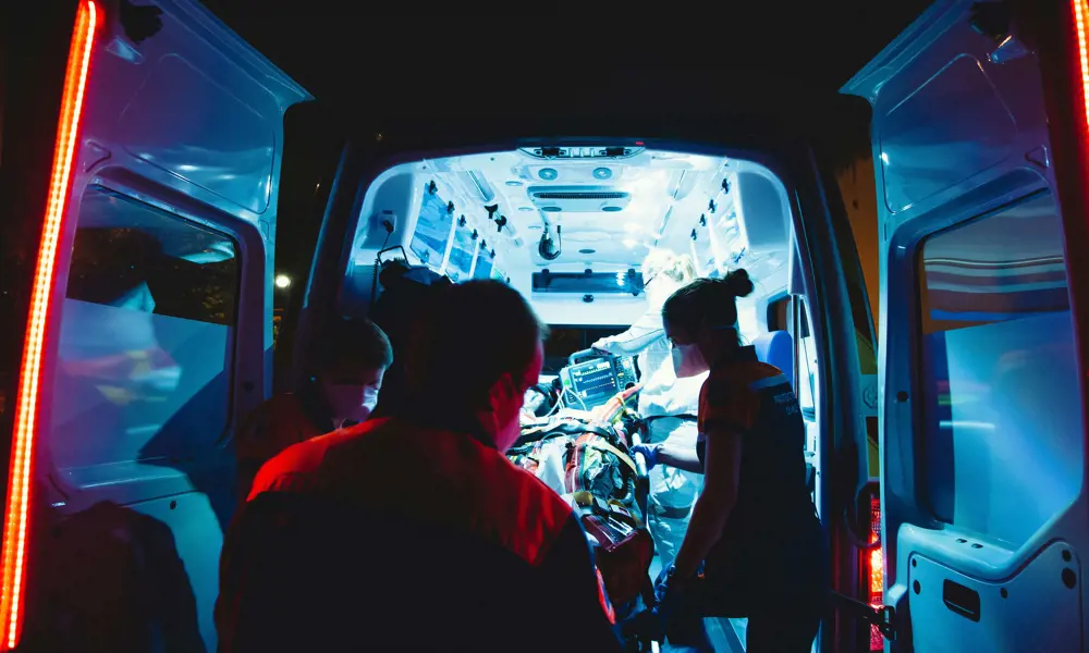 Emergency response professionals wearing masks loading the back of an ambulance with a stretcher at night.