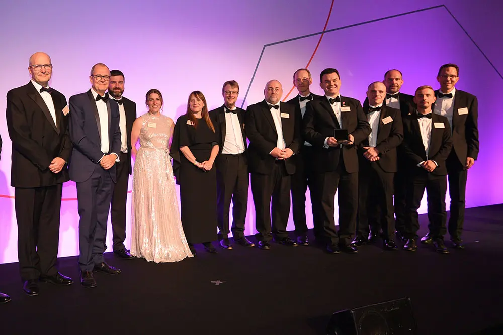 A group of people wearing formal occasionwear stand against a purple-lit background smiling at the camera