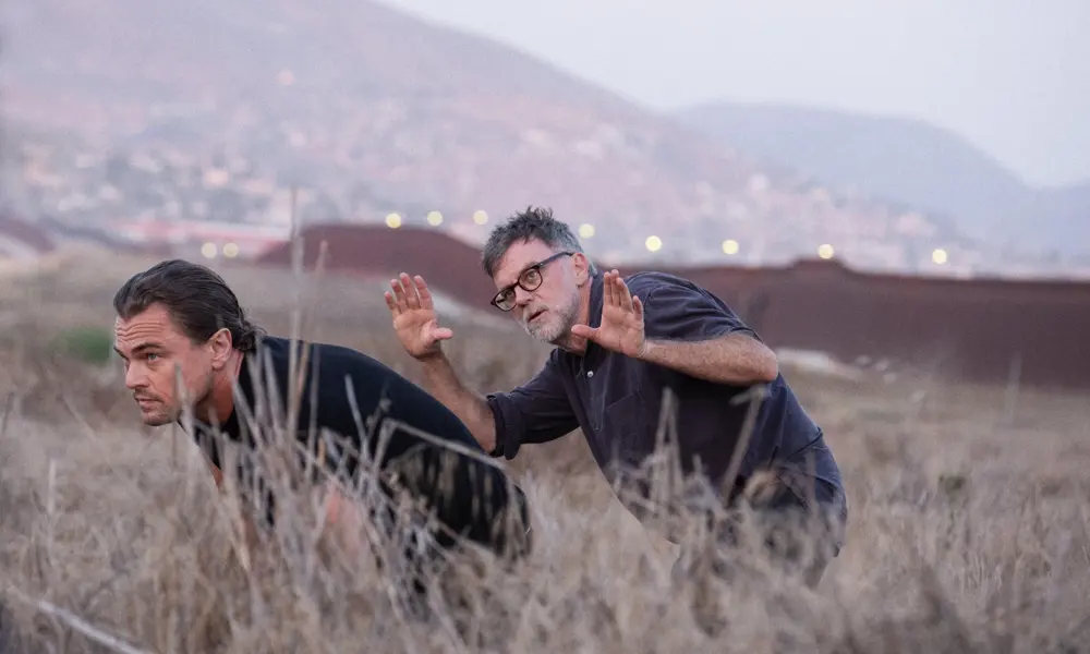 Leonardo DiCaprio and Paul Thomas Anderson are crouching in tall dry grass on a hillside, with Paul gesturing as if framing a shot, and a blurred view of mountains and a distant town in the background.