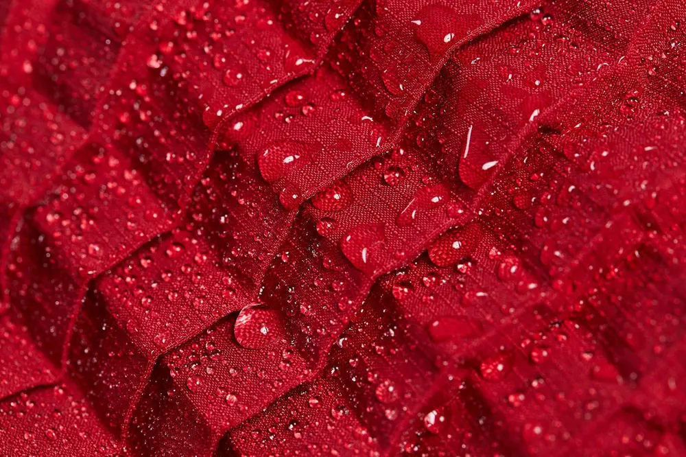A close-up of the red roof tile-like pattern that is used in petit pli's clothing with droplets of water on top of the fabric that demonstrate water resistance.