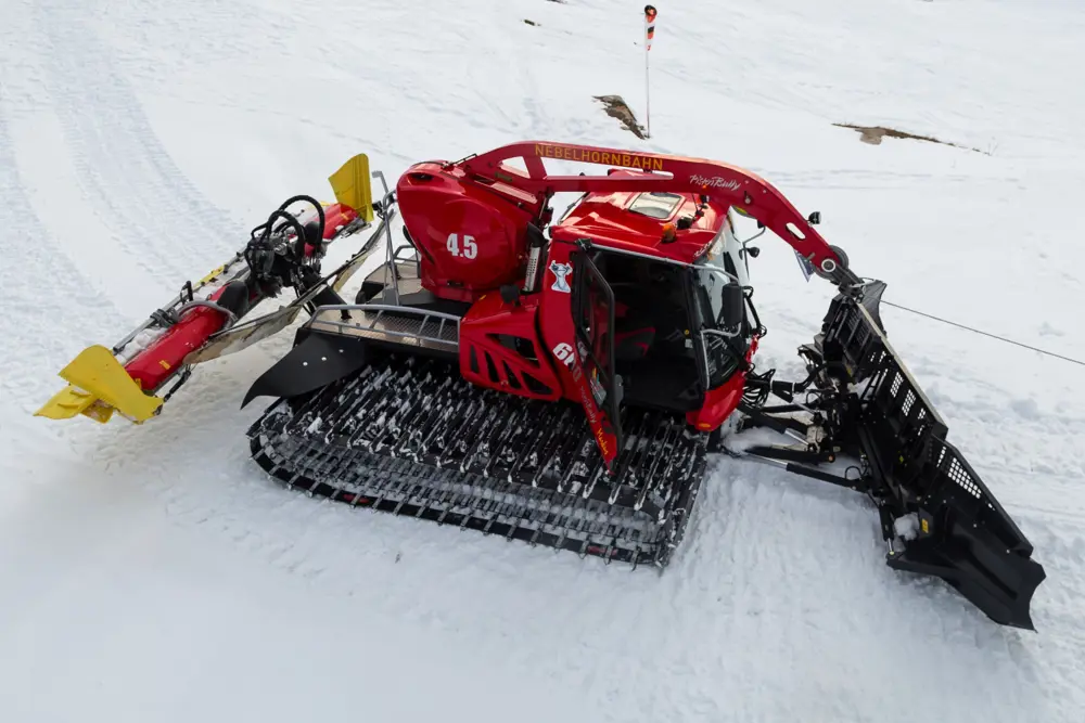 A large red snow groomer with wide tracks and a front blade is parked on a snowy slope, with its side door open and grooming equipment attached at the rear.