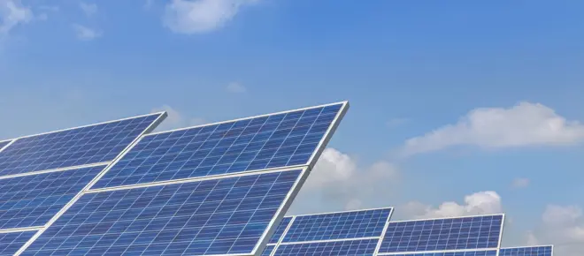 Large solar panels pictured on ground frames on a solar farm, with a blue, slightly cloudy sky above.