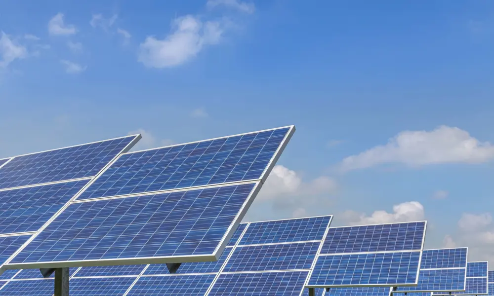Large solar panels pictured on ground frames on a solar farm, with a blue, slightly cloudy sky above.
