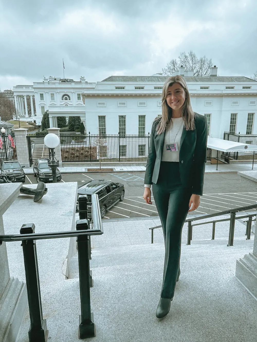 A white woman wearing a black suit and a white top stands on some steps in front of the White House in the US 