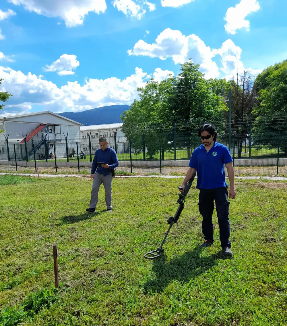 Two men walking over a field of grass, one is holding a metal detector.
