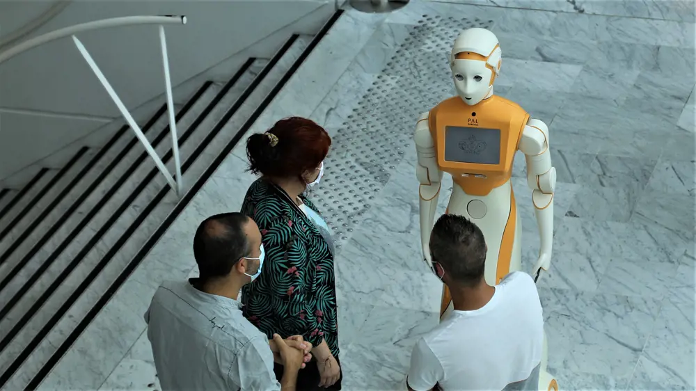 Three people stand at the top of a flight of steps, wearing covid masks and speaking to a robot with an orange torso and white arms, body and head