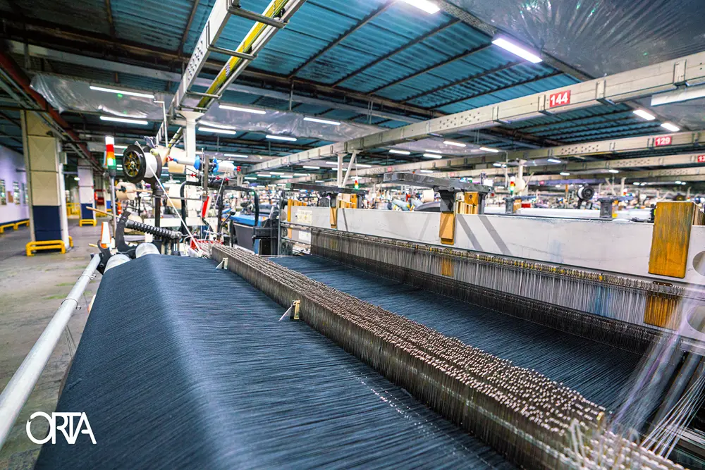 In a factory, an industrial loom is weaving denim with indigo-dyed cotton yarns, as part of the process to make jeans