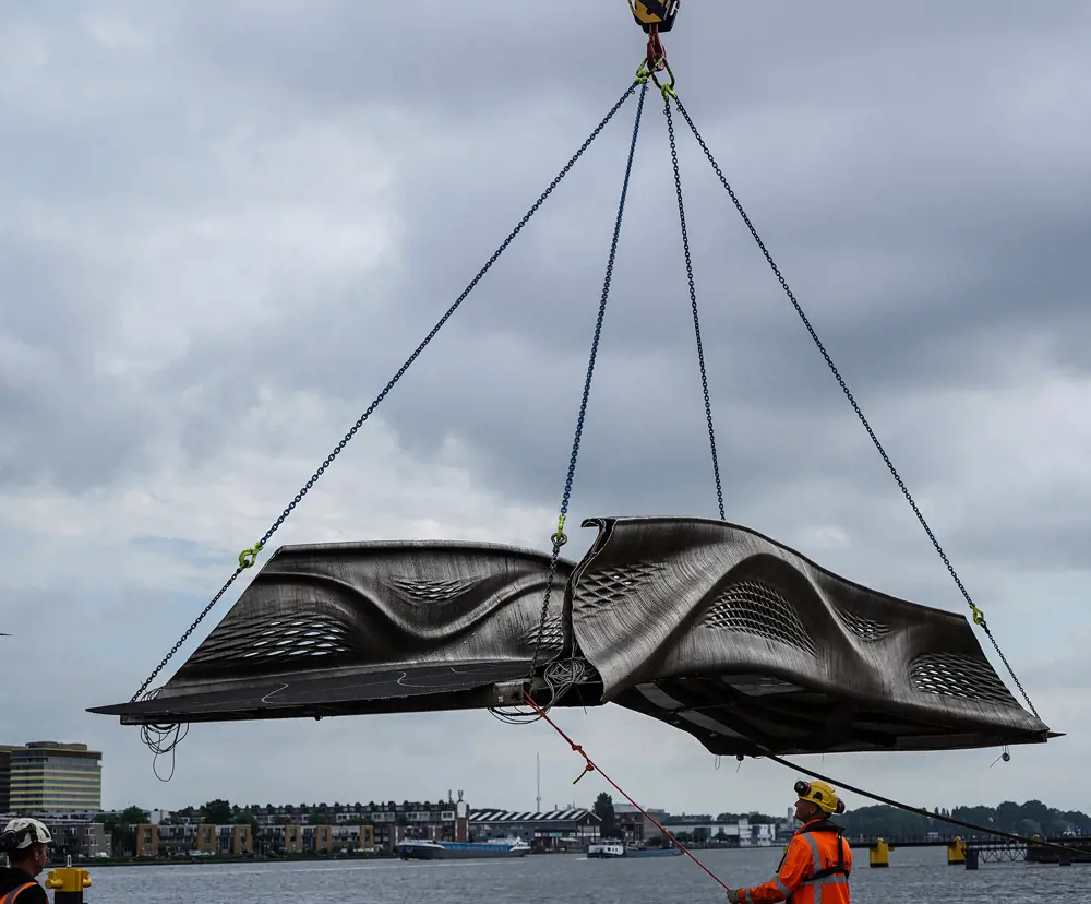 The MX3D bridge suspended from a hook in Amsterdam's main harbour area, with two men in hard hats guiding its movement.
