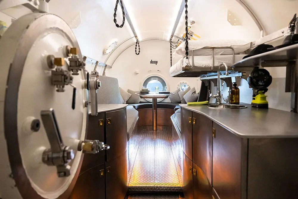 Interior of a pressurised diving chamber with metal cabinets, a small sink, bunk bed, and a seating area with cushions around a central table, seen through an open circular hatch.