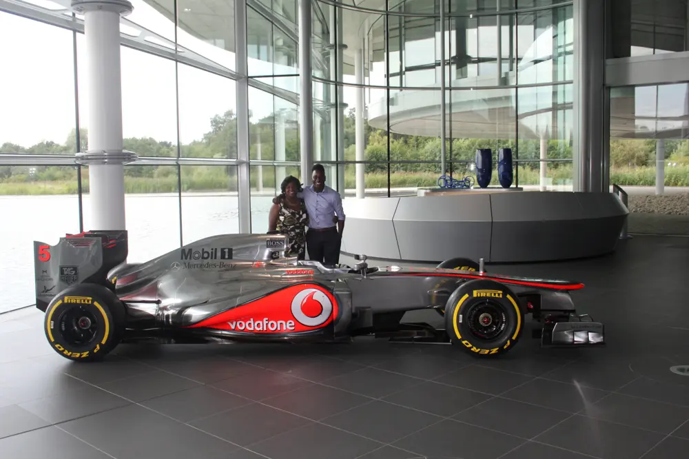 James Dornor standing with his mother, beside a silver McLaren Formula 1 car displayed inside a glass‑walled building.
