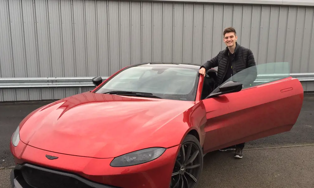 Josh Oldham smiling and standing behind an open door of a red Aston Martin.